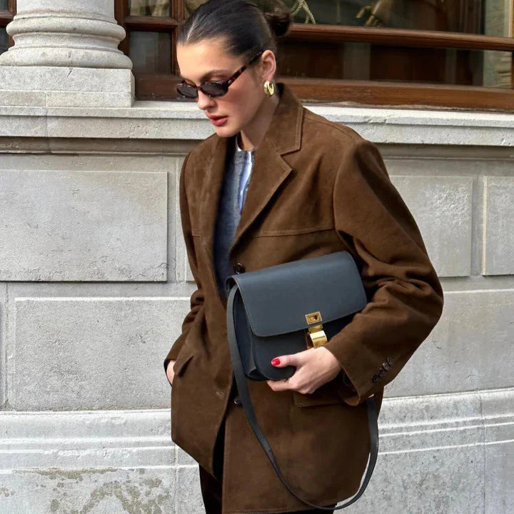 Woman in a brown coat holding a blue handbag on a city street.